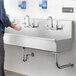 A person washing hands with soap under (5) faucets in a wall-mounted Regency hand sink.