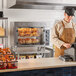 A man in a brown apron using a Cooking Performance Group countertop rotisserie oven to cook chickens.