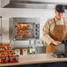 A man in a brown apron using a large metal Cooking Performance Group countertop rotisserie oven.