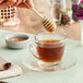 A hand using a wooden honey dipper to pour Mellody Plant-Based Vegan Honey into a glass cup of tea.