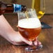 A person pouring a bottle of beer into a Stolzle Belgian beer glass on a table.