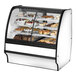 A True dual zone curved glass bakery display case with white interior and baked goods on shelves.