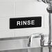 A black Lavex sign with white text that says "Rinse" above a sink.