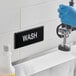 A hand in a blue glove washing a sink with a black and white Lavex Wash sign.