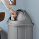 A hand throwing a crumpled paper into a Lavex Gray Half Round Trash Can with Swing Door.