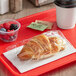 A Brioche Gourmet plant-based croissant wrapped in plastic on a red tray with a white cup of coffee and a plastic cup of berries.