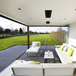 A white patio with a Bromic Heating black outdoor heater on a white surface with white and green cushions.