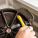 A person using a Lodge Corner / Groove scrub brush with a yellow and black handle to clean a metal pan.