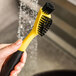 A hand holding a black and yellow Lodge Corner / Groove Scrub Brush with bristles.