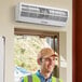 A man wearing a yellow vest and hard hat standing next to a white rectangular air curtain above a door.