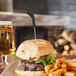 A hamburger with a bamboo cocktail pick shaped like a hockey stick inserted into the top bun, served with fries and a glass of beer in the background.