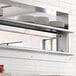 A ServIt strip warmer mounted to a stainless steel shelf above plates on a counter in a professional kitchen.