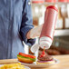 A hand holding a Vollrath Traex squeeze bottle pouring ketchup onto a burger.