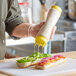 A hand holding a Vollrath Traex clear squeeze bottle with yellow cap and base, pouring mayonnaise on a sandwich.