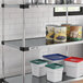 A Regency galvanized steel shelf holding white containers with green and red lids and cans of mushrooms.