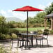A black table and chairs under a Lancaster Table & Seating red umbrella on a stone patio.