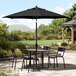 A black table and chairs under a black Lancaster Table & Seating umbrella on a patio.