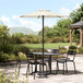 A round ivory table with black chairs and a Lancaster Table & Seating black umbrella on a stone patio.