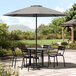 A Lancaster Table & Seating pewter gray umbrella over a table and chairs on a stone surface.