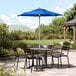 A table with black chairs and a Lancaster Table & Seating blue umbrella on a stone patio.