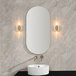 A white wall-mounted vanity light with cylindrical glass shades over a rectangular mirror and sink.