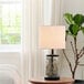 A Globe farmhouse table lamp with a fillable watered glass base on a wooden table with a plant inside.