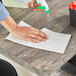 A person cleaning a table with a white WypAll heavy-duty wiper and a spray bottle.