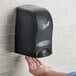 A person using a Scott black manual liquid hand soap dispenser on a wall.