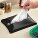 A hand using a Tork Xpressnap black napkin dispenser on a counter.