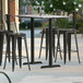 A Lancaster Table & Seating bar height table with four black bar stools on a patio.