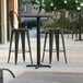 A Lancaster Table & Seating bar height table with two black outdoor cafe barstools on a patio.