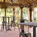 A Lancaster Table & Seating Paladina bar height table with 2 black outdoor cafe barstools on a covered patio.