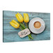 A white canvas print of yellow tulips and a coffee cup on a blue wood table.