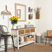 A BrandtWorks natural grain finish mirror in a white room over a table with a bouquet of yellow flowers and a white chair with a blanket and a basket with a blanket inside.