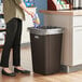 A woman wearing black pants puts a plastic bag in a brown Lavex square trash can.