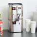 A Grindmaster stainless steel coffee shuttle and warmer on a counter next to cups.
