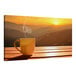 A coffee cup on a table with a view of a sunset over mountains.