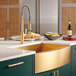 A Ruvati Terraza stainless steel farmhouse sink with brass tone accents in a kitchen with green cabinets.