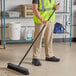 A man in a safety vest using a Lavex polypropylene push broom with flagged bristles and a metal handle to sweep a grocery store aisle.