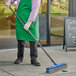 A man in a green apron using a Lavex blue push broom with gray flagged bristles to sweep the sidewalk.