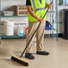 A man in a safety vest using a Lavex wood push broom with unflagged bristles to sweep a grocery store aisle.