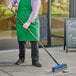 A man in a green apron using a Lavex blue push broom with gray flagged bristles to sweep the sidewalk.
