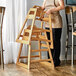 A woman standing next to a Lancaster Table & Seating wooden high chair with a natural finish.