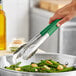A hand using Fourt&#233; green stainless steel scalloped tongs to serve broccoli from a bowl.
