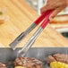 A person using Fourt&#233; stainless steel tongs with red coated handles to grill meat.