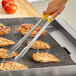 A person using yellow Fourt&#233; stainless steel tongs to cook meat on a grill.