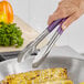 A person using Fourté stainless steel scalloped tongs with purple handles to hold corn over a pan.