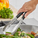 A person using Fourt&#233; black stainless steel tongs to serve green vegetables.