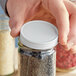 A person holding a jar with a white metal lid filled with seeds.