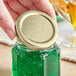 A hand holding a jar of green liquid with a gold metal lid.